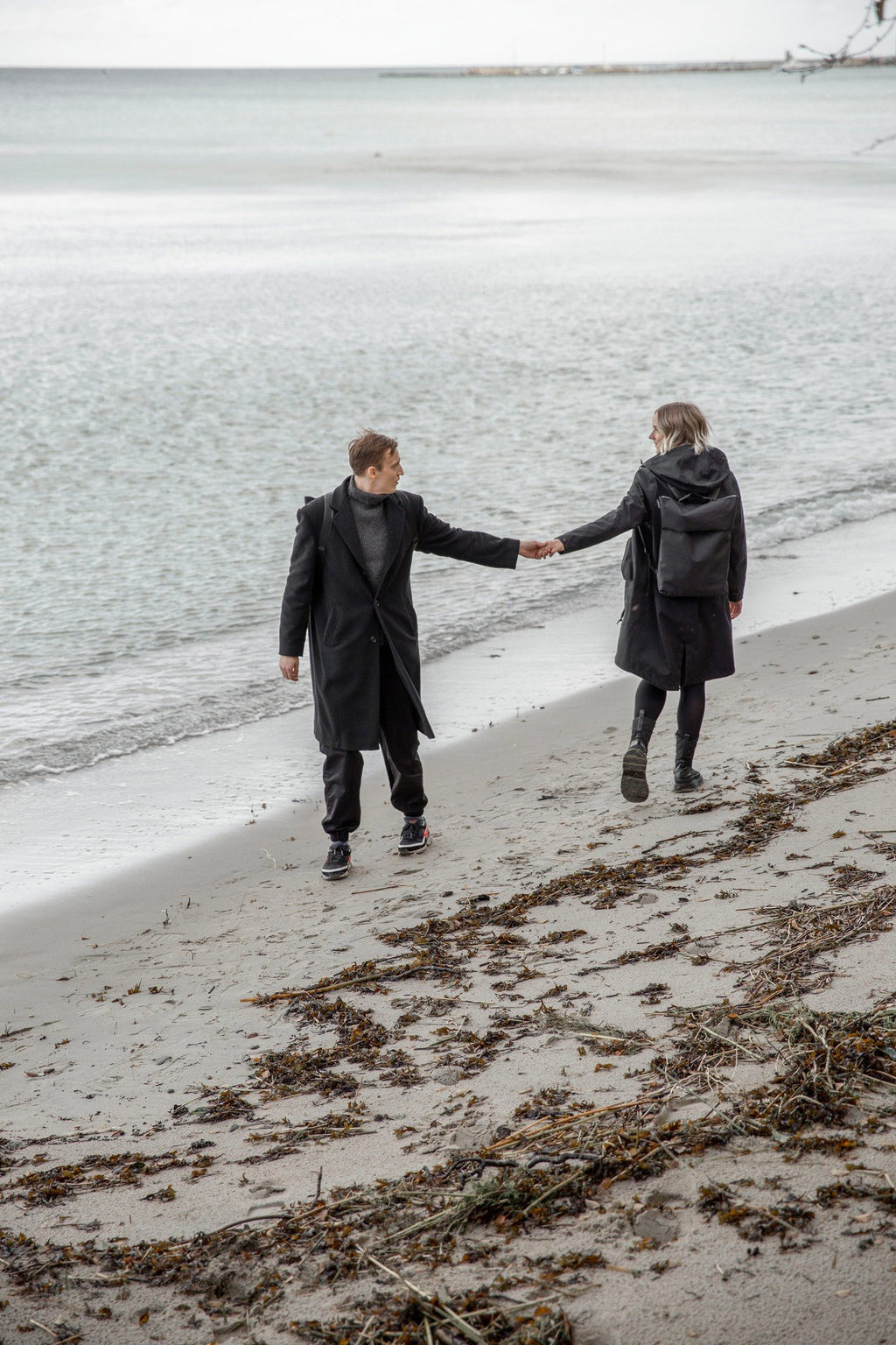 2. Unisex Nordhale HØLAR Weatherproof Backpack worn by male and female models walking on a beach, highlighting outdoor versatility