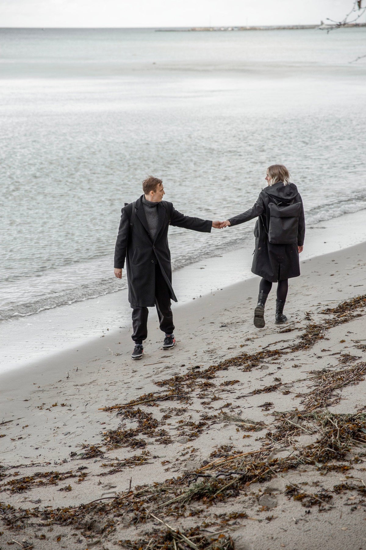 2. Unisex Nordhale HØLAR Weatherproof Backpack worn by male and female models walking on a beach, highlighting outdoor versatility