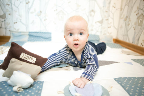 1. Baby playing with Karloova Pillow Heap on patterned blanket, holding brown pillow with 'ROBIN' label