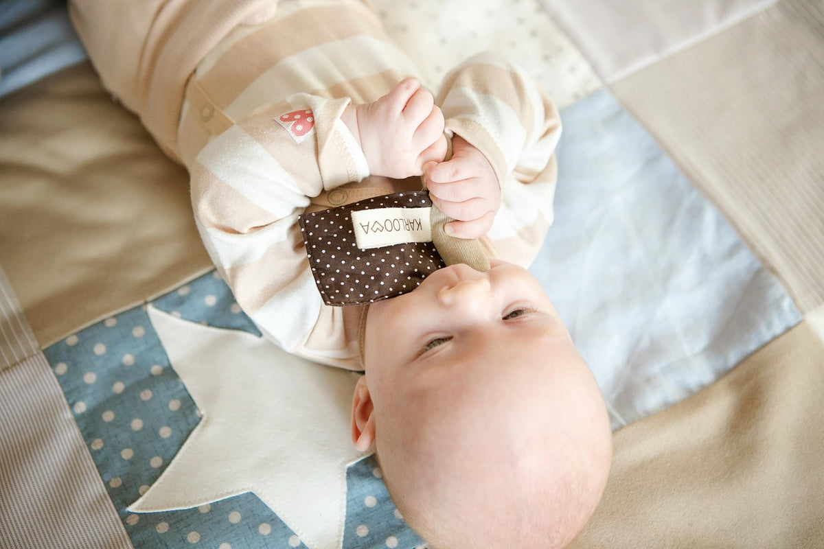 1. Baby lying on patchwork blanket holding Karloova textile flag toy with brown polka dots