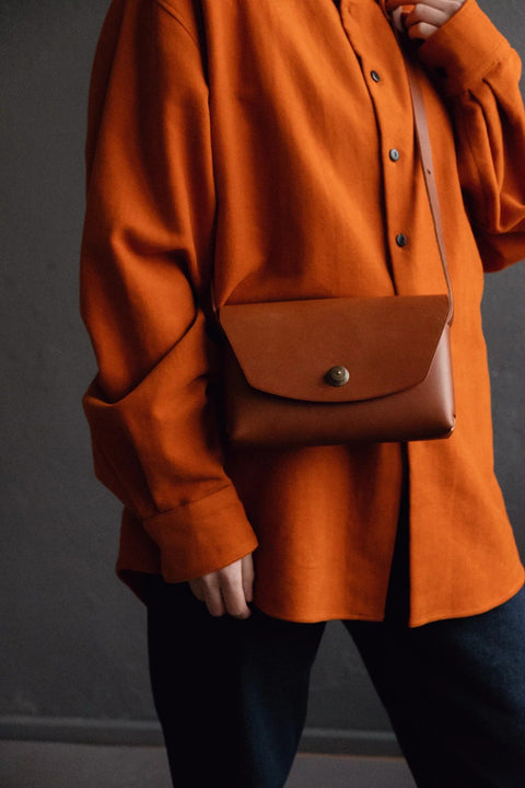 1. Women's rust brown leather clutch bag by Mirgoods, worn crossbody with orange shirt in lifestyle setting