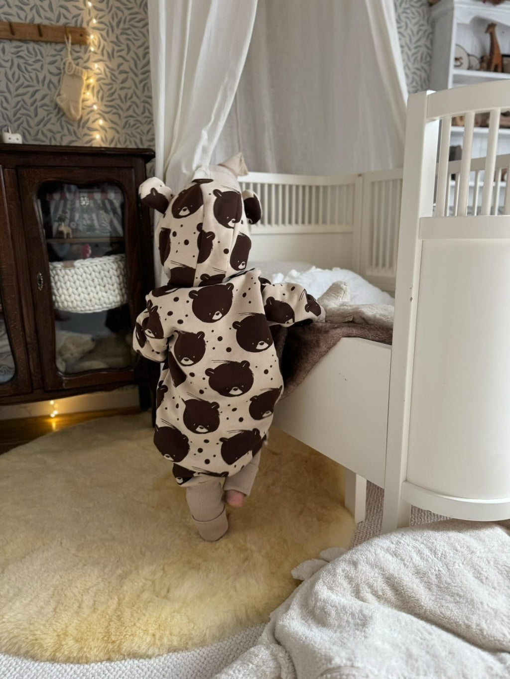 1. Back view of baby in beige bear-patterned jumpsuit with hood in a nursery setting
