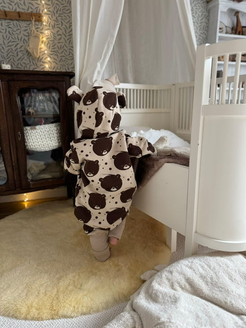 1. Back view of baby in beige bear-patterned jumpsuit with hood in a nursery setting