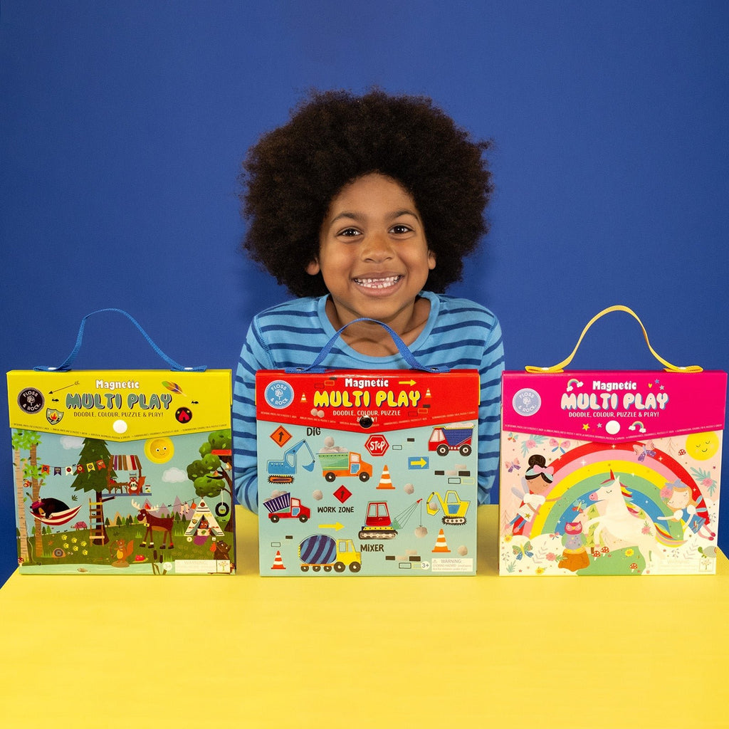 1. Child smiling with three Floss & Rock Magnetic Multi Play Sets on a yellow table against blue background