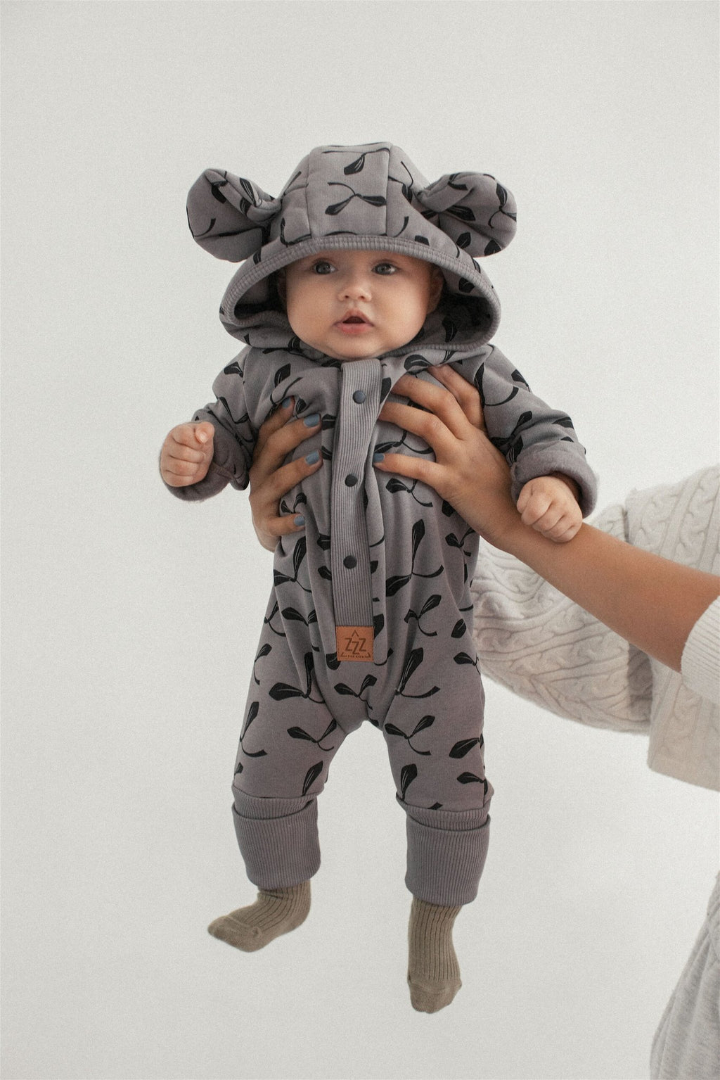 1. Baby wearing steel jumpsuit with maple seed print and hood with ears, held up by adult hands