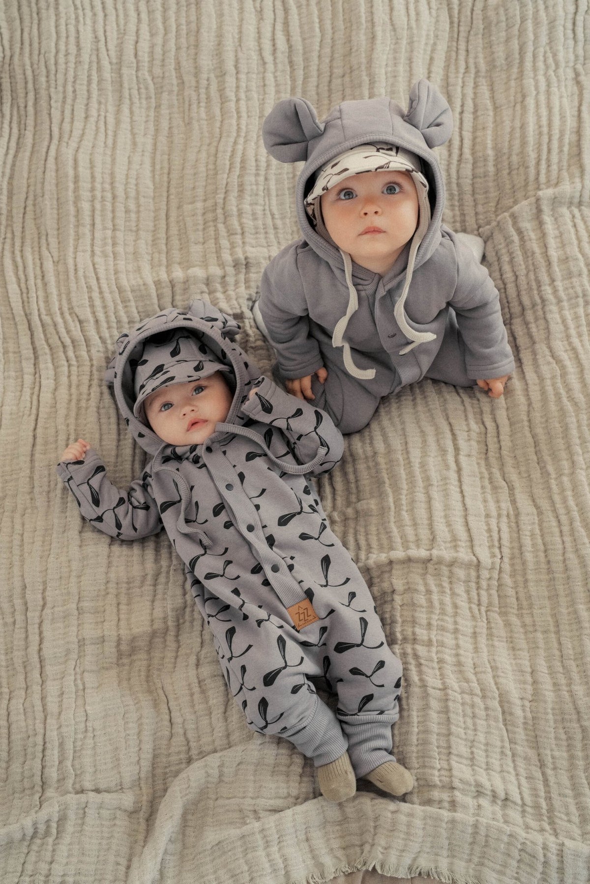 1. Two babies wearing steel jumpsuits with maple seed print and hood with ears, lying on a beige blanket