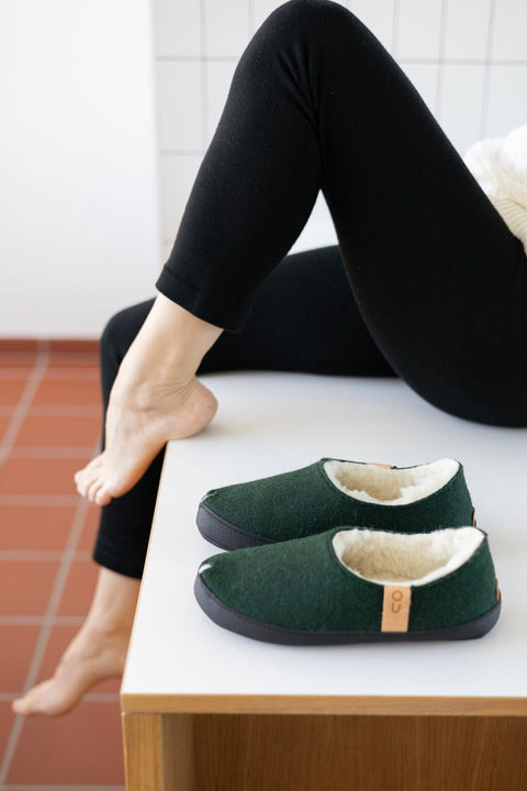 1. Unisex dark green TOKU Budapest slippers with lambswool lining, shown in lifestyle setting with model lounging