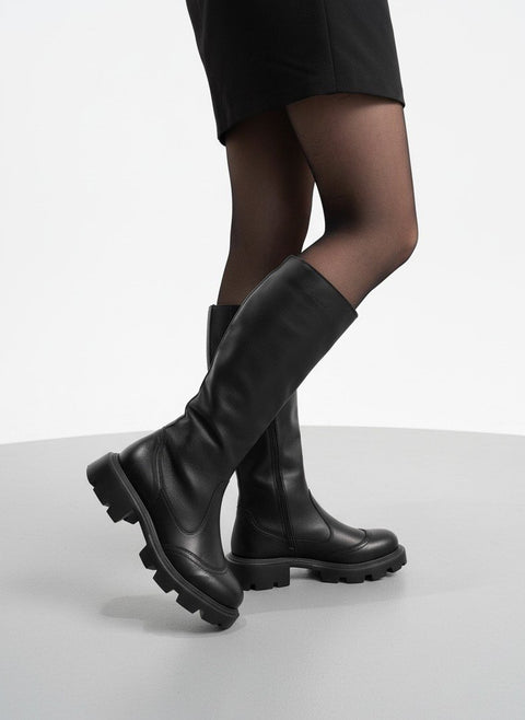 1. Woman wearing black high nappa leather boots with chunky sole, standing on a light grey surface