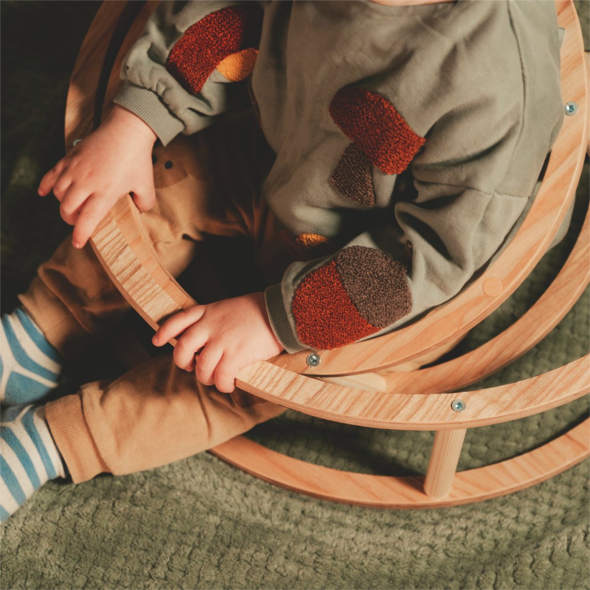 5. Child sitting inside large wooden ring on green textured surface, wearing colorful patchwork sweater