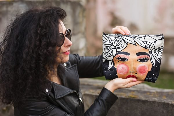 1. Woman holding Garbanota YOKO makeup bag in black with artistic face print, outdoor setting