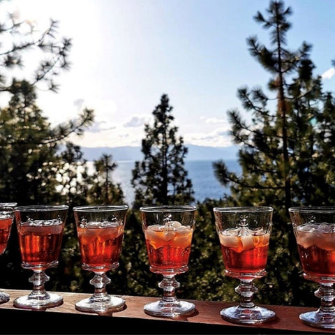 2. Six La Rochère Bee glasses filled with red drinks on a balcony with a scenic forest and lake view
