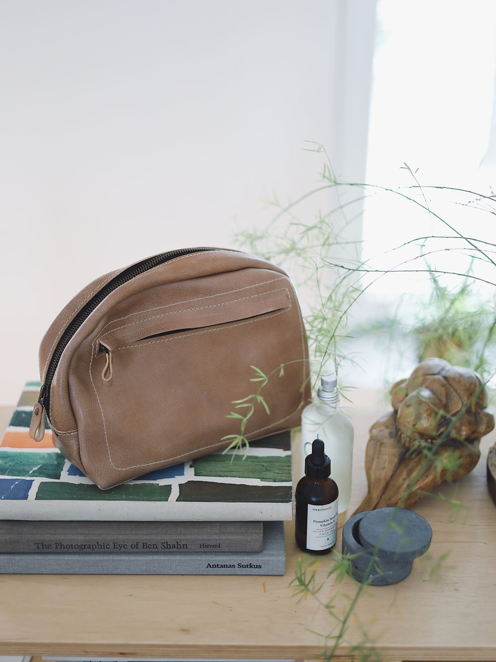 1. Beige DOUGLAS Toiletry Bag by TOKS VISOKS on a wooden table with books and plants, showcasing eco-friendly design