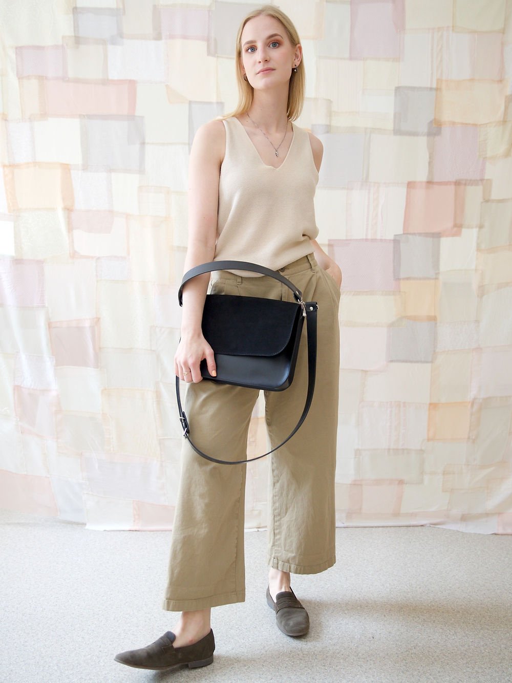 14. Woman holding TOKS VISOKS ASTORIA black suede handbag, styled with beige top and khaki pants