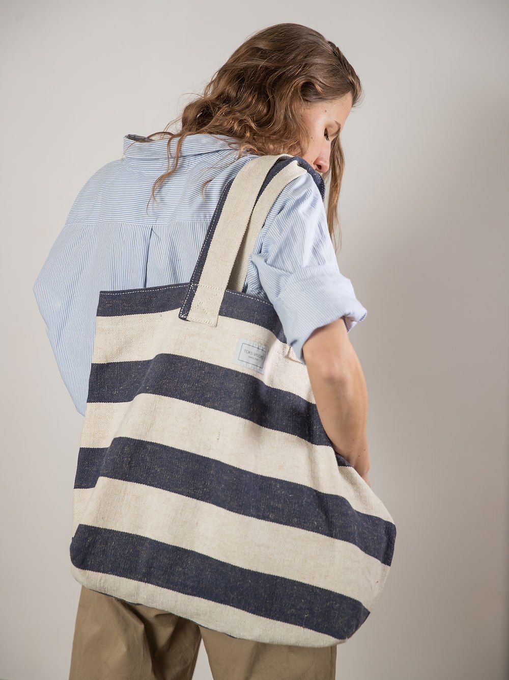 11. Woman showing back view of TOKS VISOKS oversized beach bag in reclaimed stripe canvas, studio setting