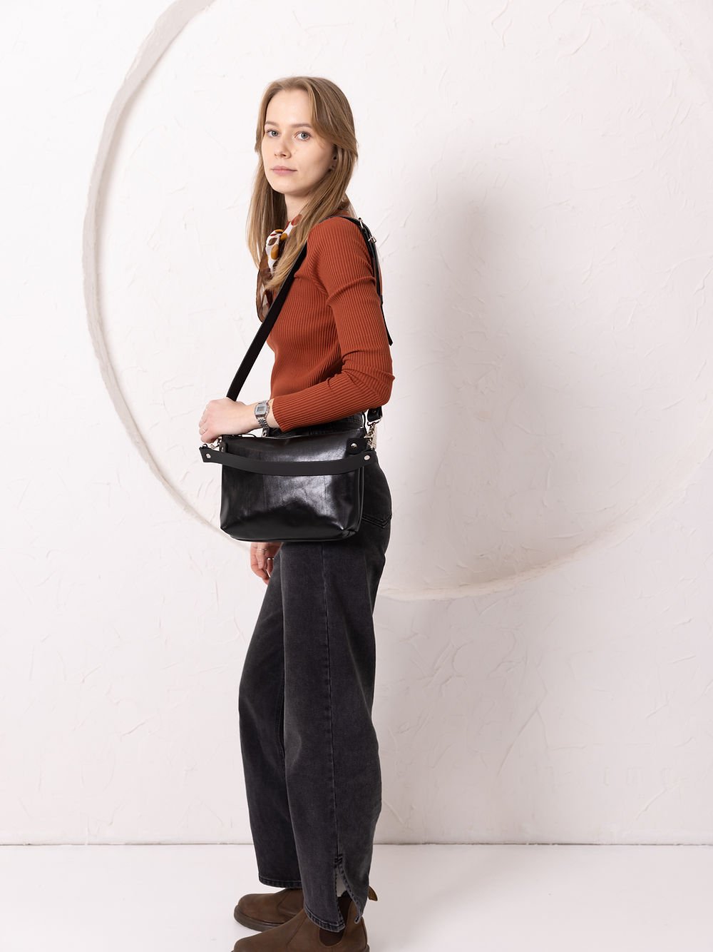 16. Women's black TOMMY leather bag by TOKS VISOKS worn on shoulder, side view in studio setting