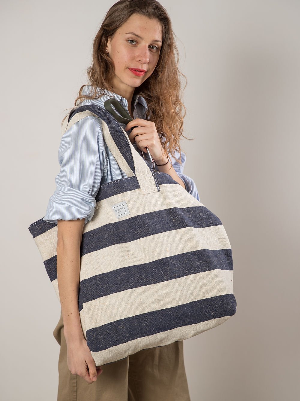 8. Woman posing with TOKS VISOKS oversized beach bag in reclaimed stripe canvas, studio setting
