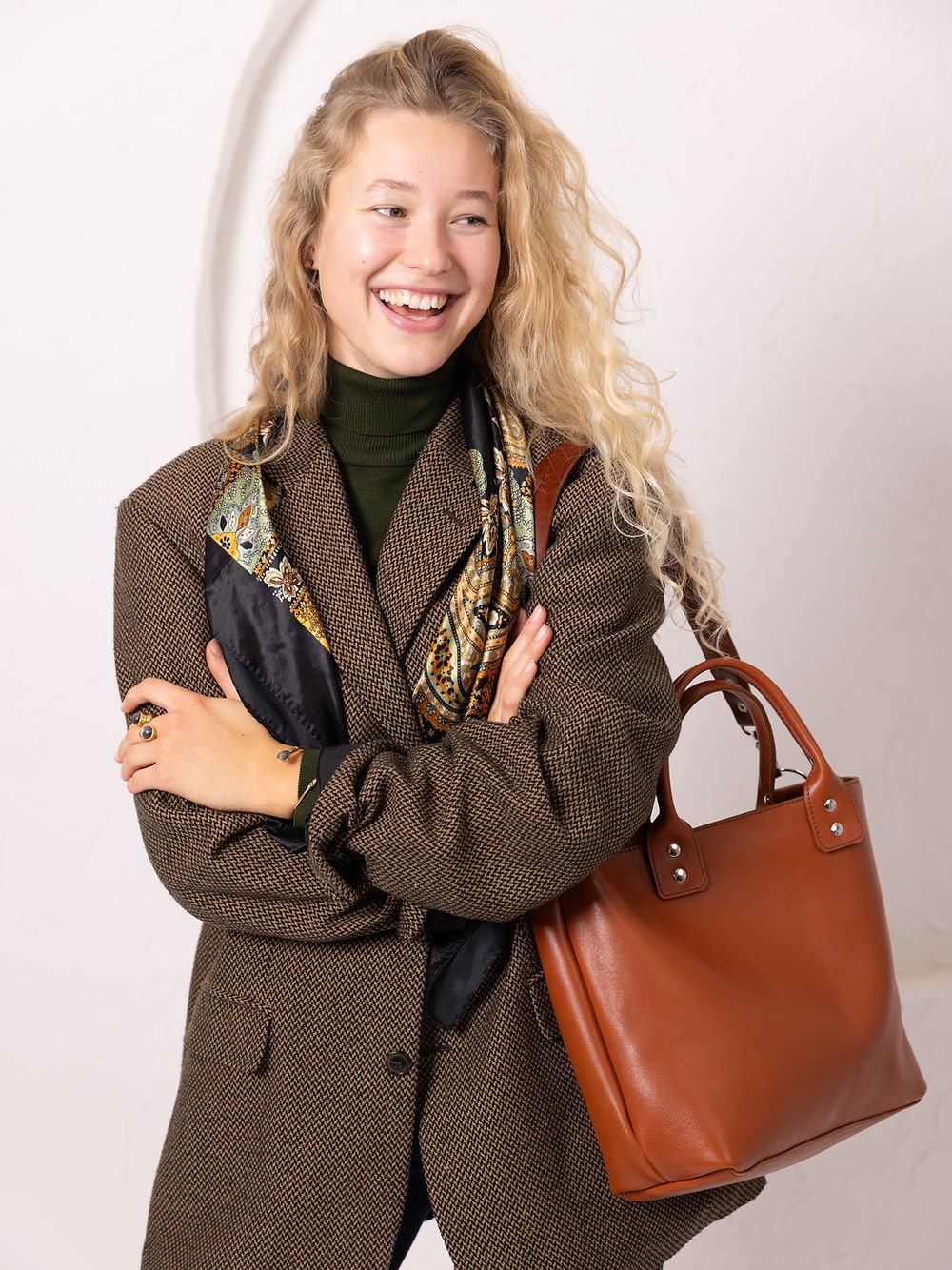 2. Woman smiling with TOKS VISOKS MIGLE leather handbag in ginger, styled with a scarf