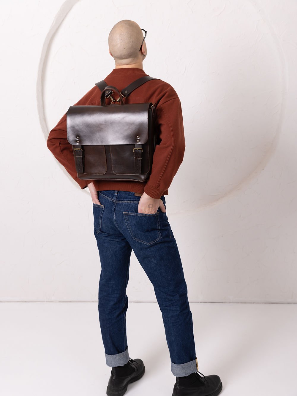 4. Male model adjusting TOKS VISOKS BENJAMIN Backpack in dark chocolate leather, highlighting front pockets and buckle