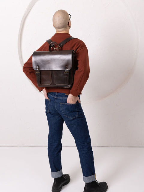 4. Male model adjusting TOKS VISOKS BENJAMIN Backpack in dark chocolate leather, highlighting front pockets and buckle