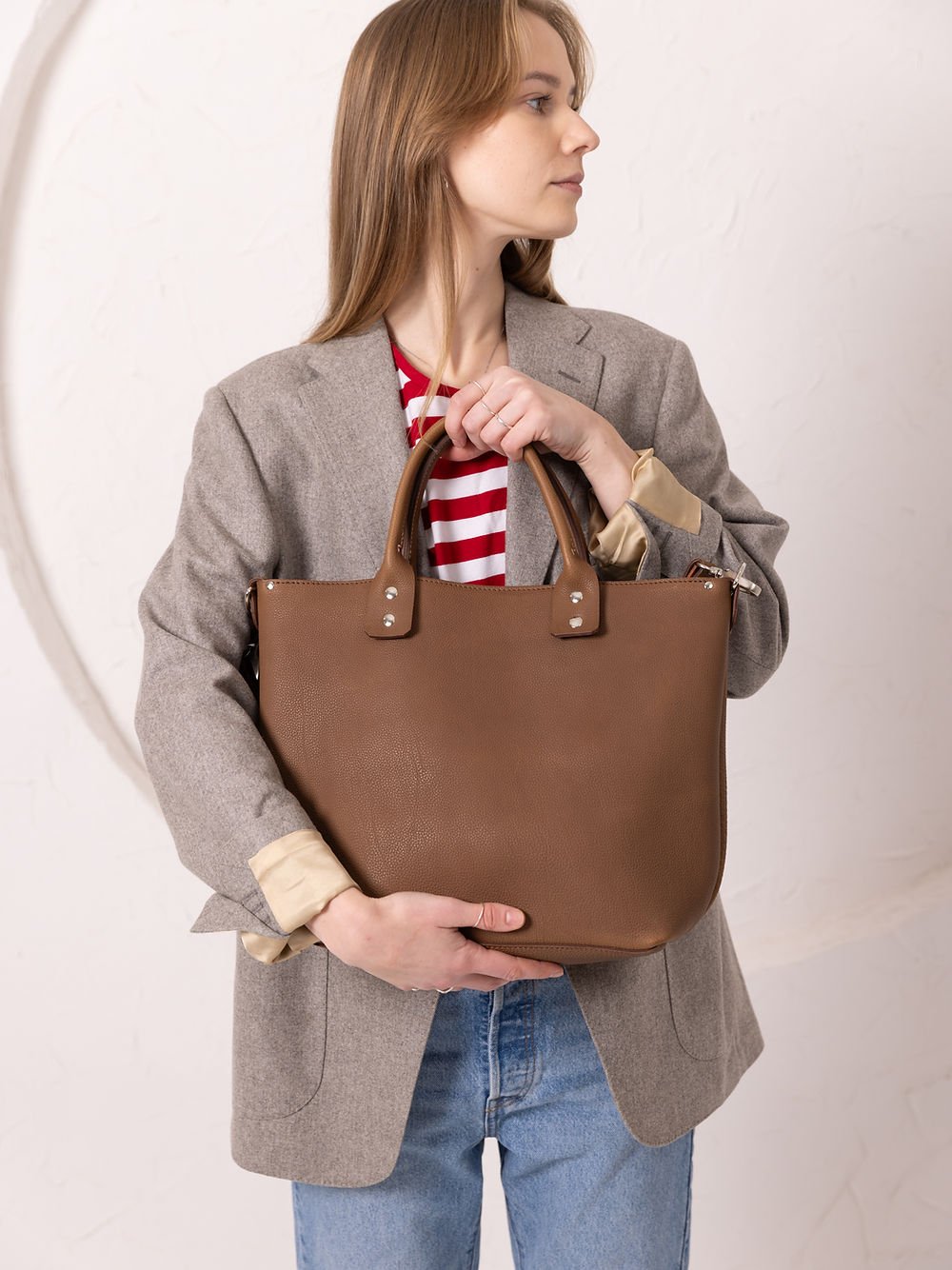 1. Woman holding MIGLE Leather Handbag in cappuccino by TOKS VISOKS, styled with a blazer and jeans