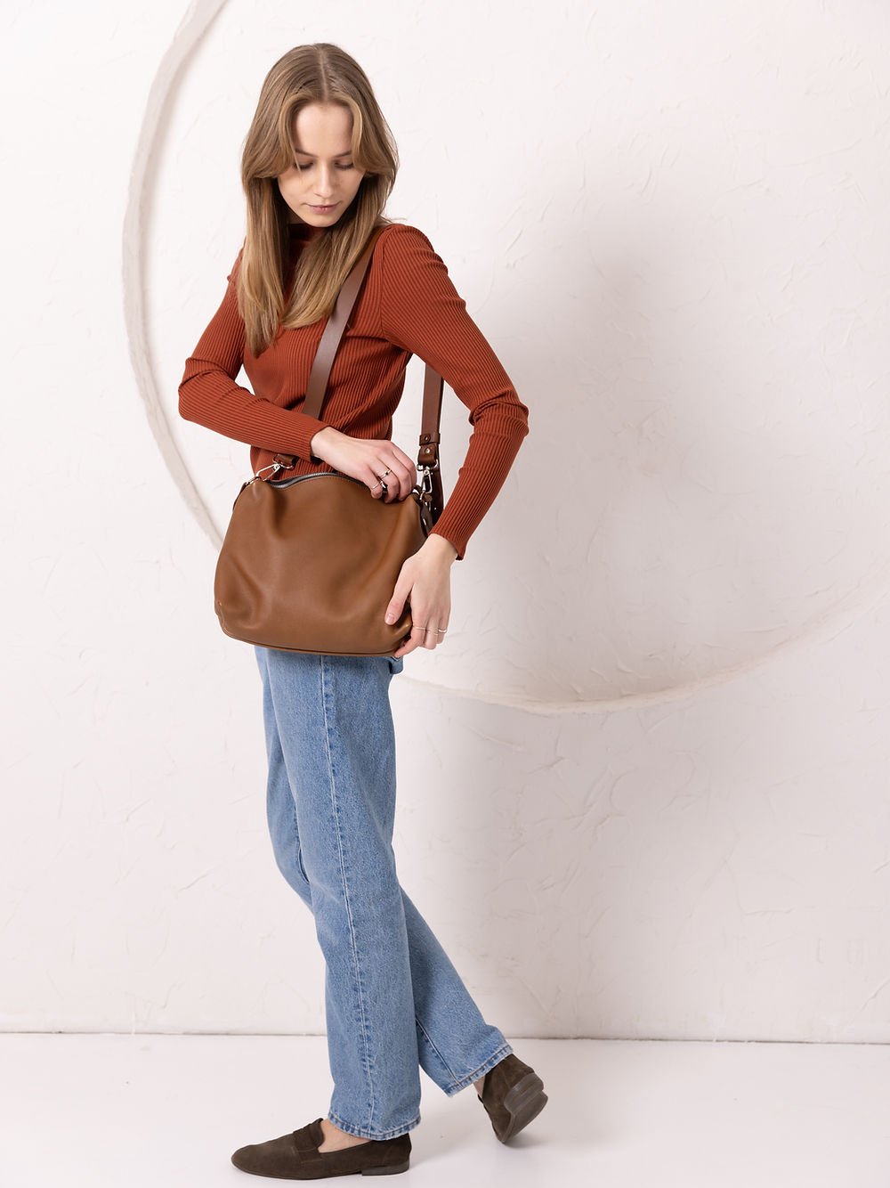 4. Woman adjusting TOKS VISOKS ROBIN leather handbag in camel brown, studio setting