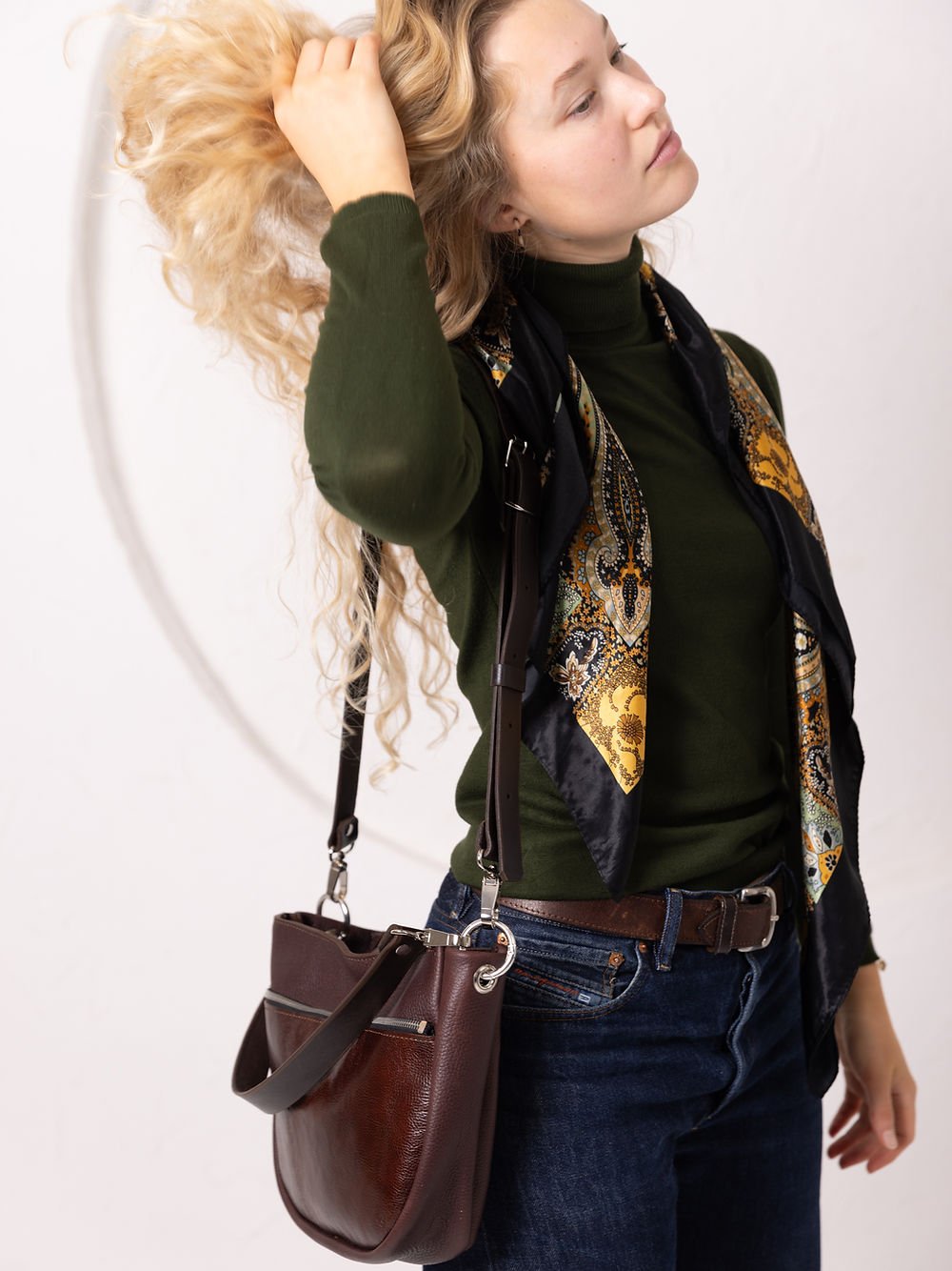 7. Woman adjusting hair while wearing TOKS VISOKS STELLA leather handbag in brown and cognac, styled with scarf in studio