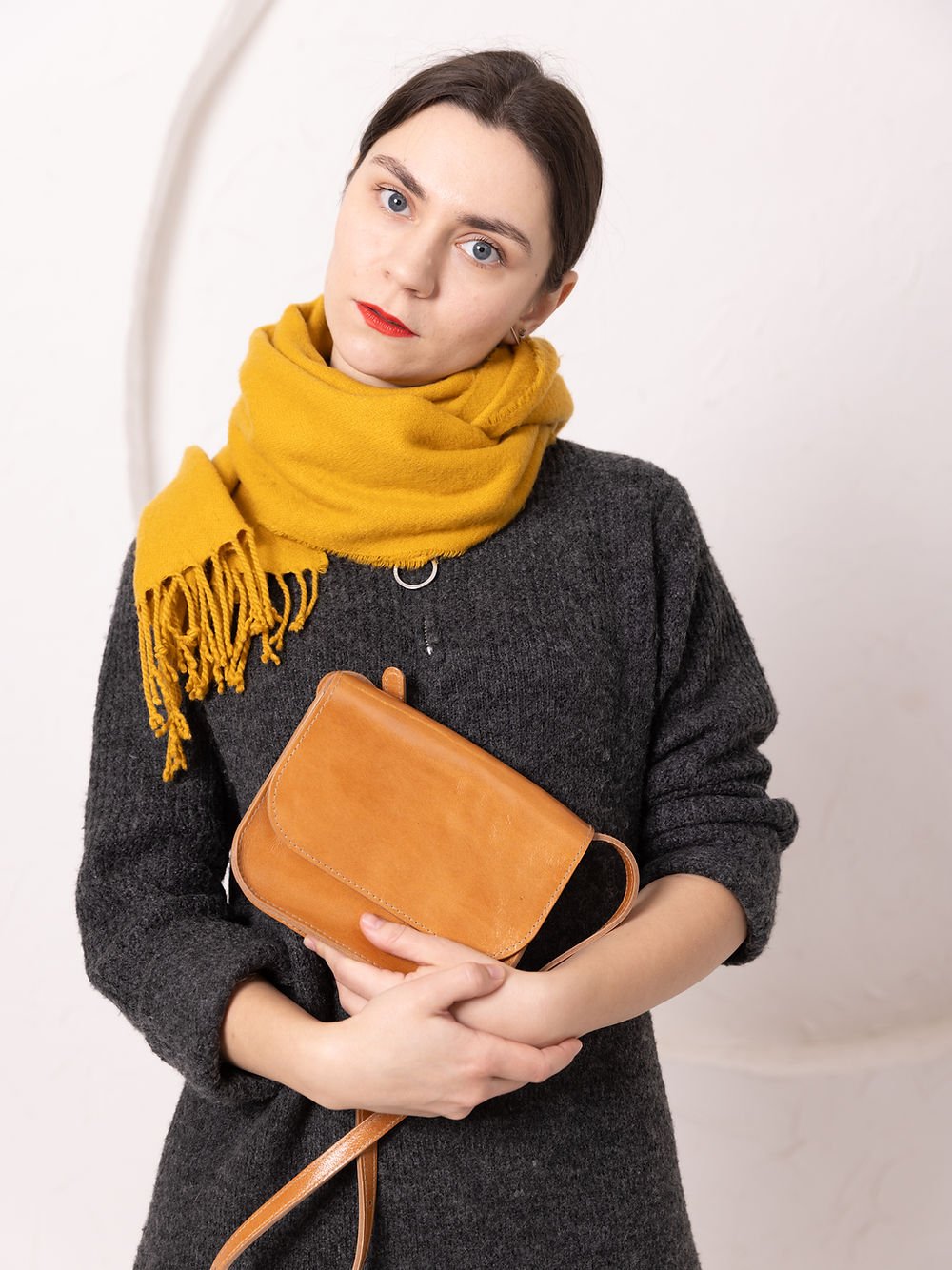 1. Woman holding TOKS VISOKS RUBY Purse in caramel leather, styled with a yellow scarf and dark sweater