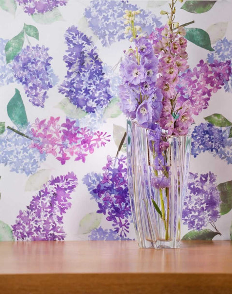 1. Lilac garden wallpaper in light purple with watercolor floral design behind a clear vase of purple flowers on a wooden surface