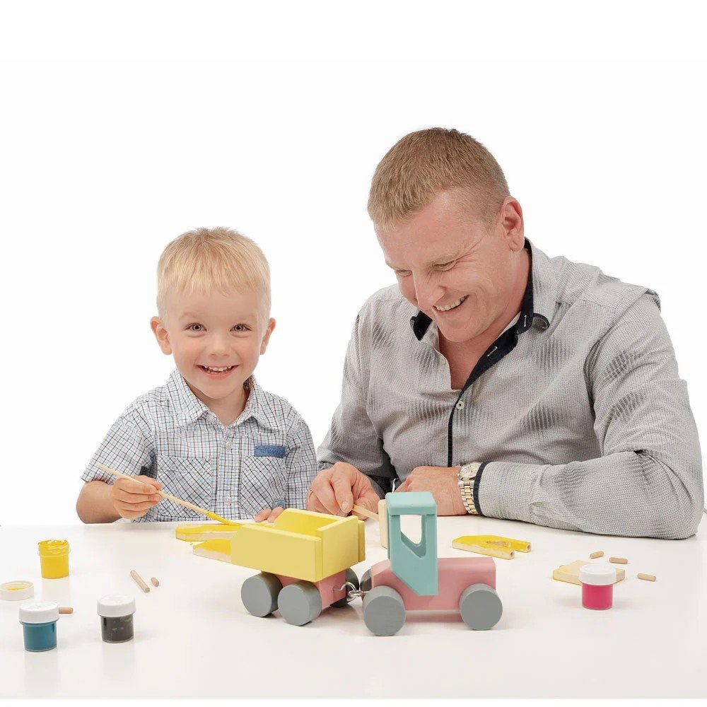 1. Smiling child and adult painting a wooden toy tractor with yellow and pink watercolors, highlighting the interactive DIY aspect