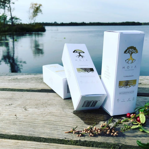 2. HOIA homespa Sacred Forest Peat Mask boxes on wooden deck by a lake, showcasing natural and eco-friendly packaging