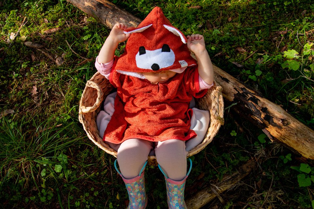 3. Toddler sitting in basket wearing RÄTT Short-Sleeve Poncho FOX, surrounded by nature