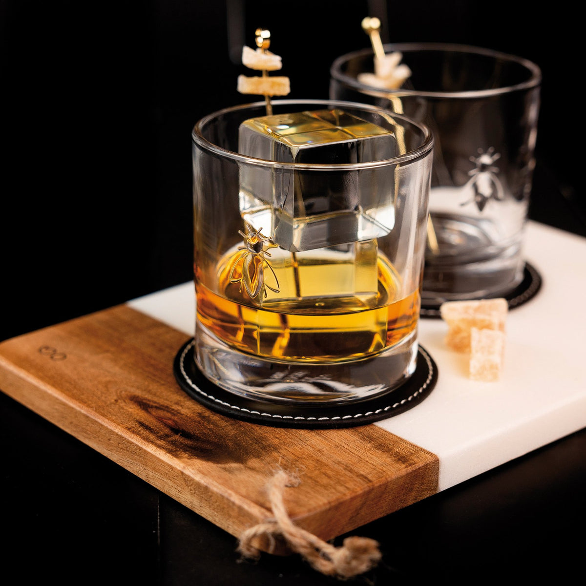 1. Two La Rochère Bee Whiskey Glasses with embossed bee design on a wooden board, filled with whiskey and ice cubes, creating a stylish and elegant setting