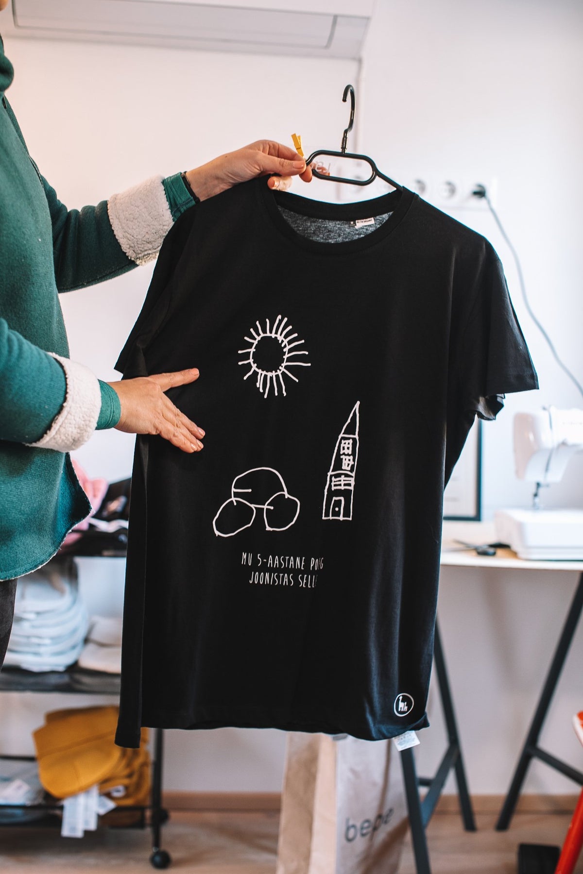 1. Black Giraffe Brand black T-shirt with child's drawing of sun and building, held by a person in a casual setting