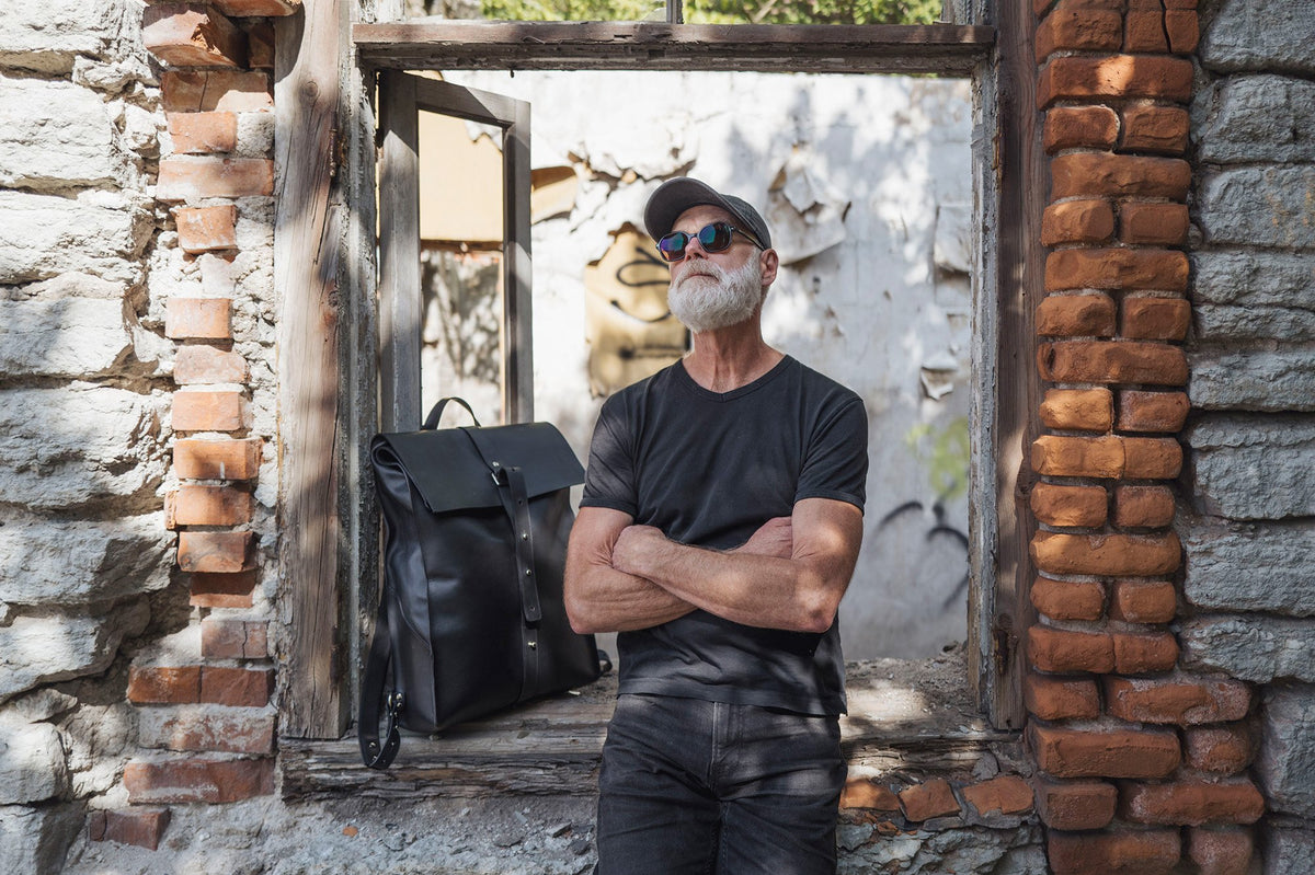 1. Man in black t-shirt and cap standing by a window with a black leather knapsack on ledge, urban setting
