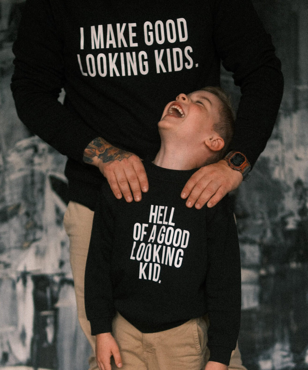 1. Dad and child wearing Black Giraffe Brand matching black sweatshirts with playful text, dad's reads 'I Make Good Looking Kids' and child's reads 'Hell of a Good Looking Kid', in a cozy setting
