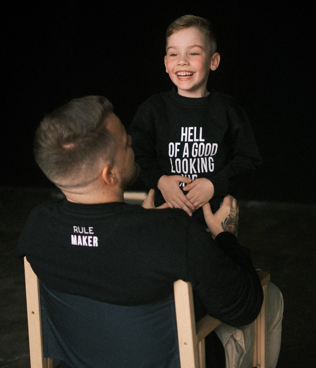 6. Father and son in Black Giraffe Brand matching black sweatshirts, dad's with 'Rule Maker' on the back and child's with 'Hell of a Good Looking Kid', sharing a playful moment