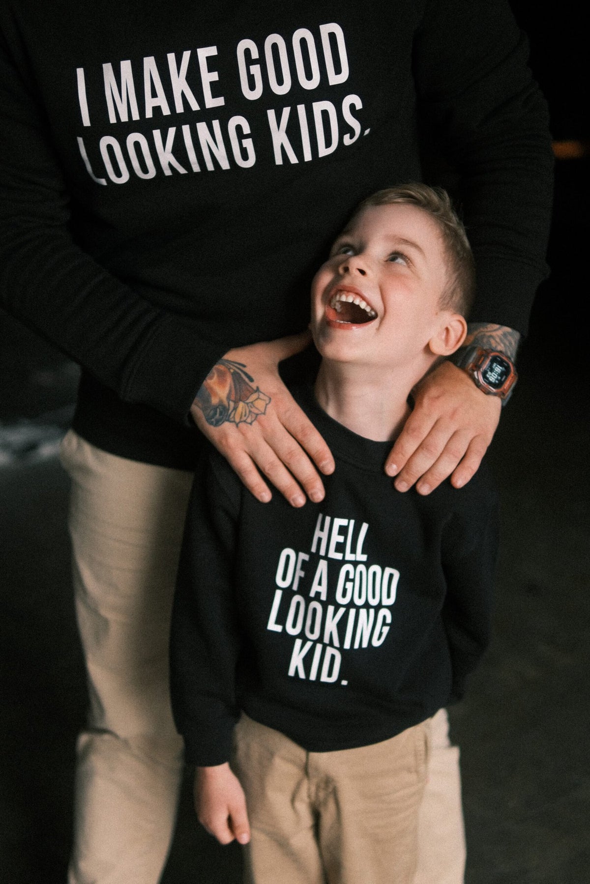 2. Father and son in Black Giraffe Brand matching black sweatshirts, dad's with 'I Make Good Looking Kids' and child's with 'Hell of a Good Looking Kid', sharing a joyful moment