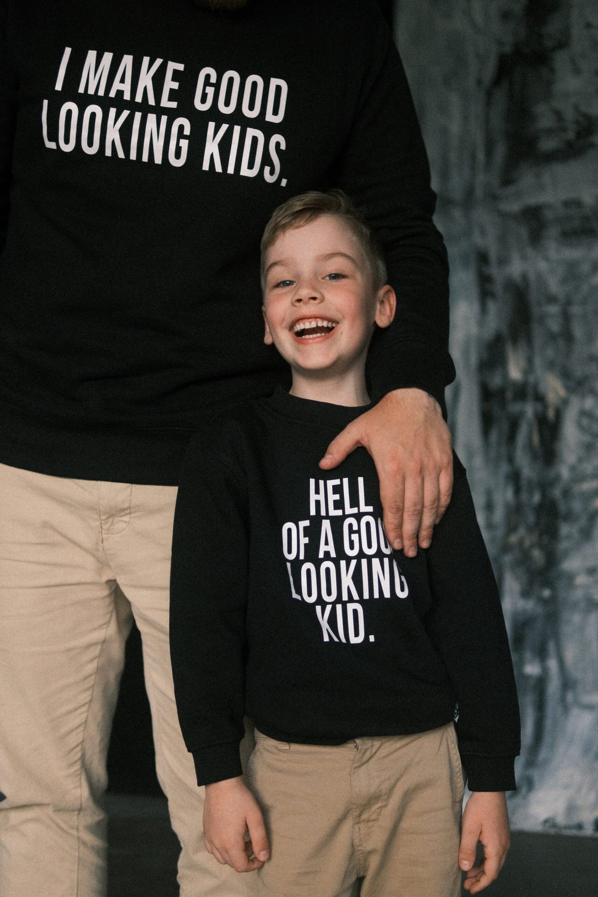 3. Black Giraffe Brand matching black sweatshirts for dad and child, featuring 'I Make Good Looking Kids' and 'Hell of a Good Looking Kid' text, shown in a warm family setting