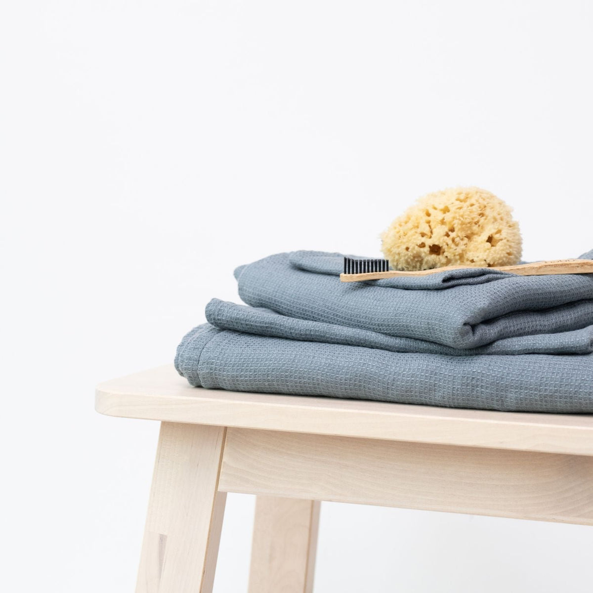 6. Stack of blue fog waffle linen towels on a stool with a natural sponge and bamboo toothbrush