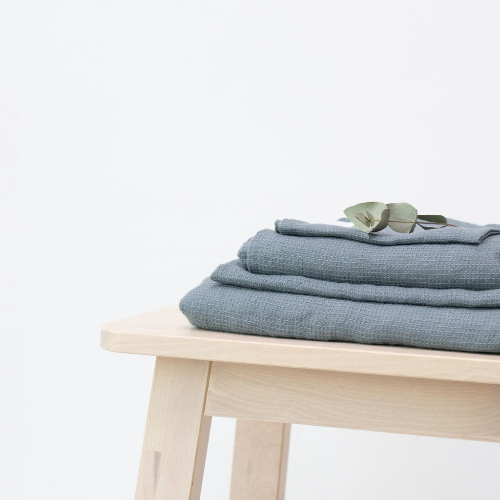 1. Set of blue fog waffle linen towels stacked on a light wooden stool with a eucalyptus leaf on top