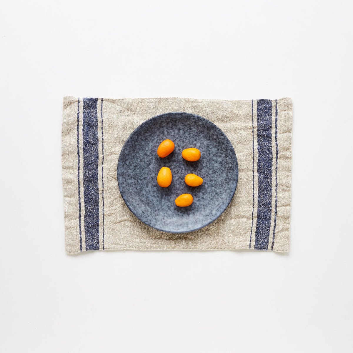 1. Blue vintage stripe linen placemat with a grey plate and kumquats on a white background