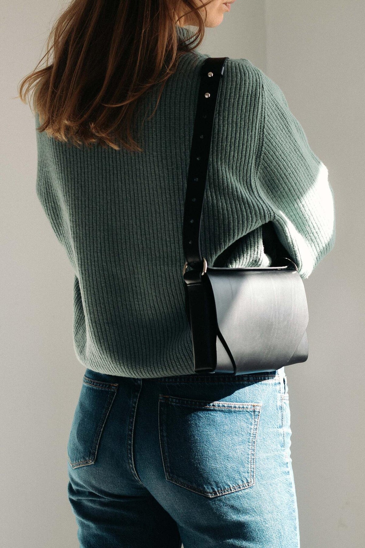1. Woman wearing black leather Stella Soomlais shoulder bag with adjustable strap over green ribbed sweater and jeans in natural light