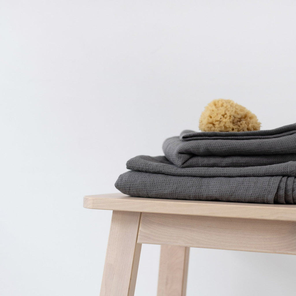 1. Stack of charcoal waffle linen towels on a wooden stool with a natural sponge on top, minimalist setting