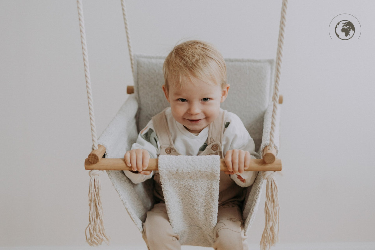 2. Child sitting in Nofi KIDS bouclé ice grey swing with oak wood poles, smiling indoors