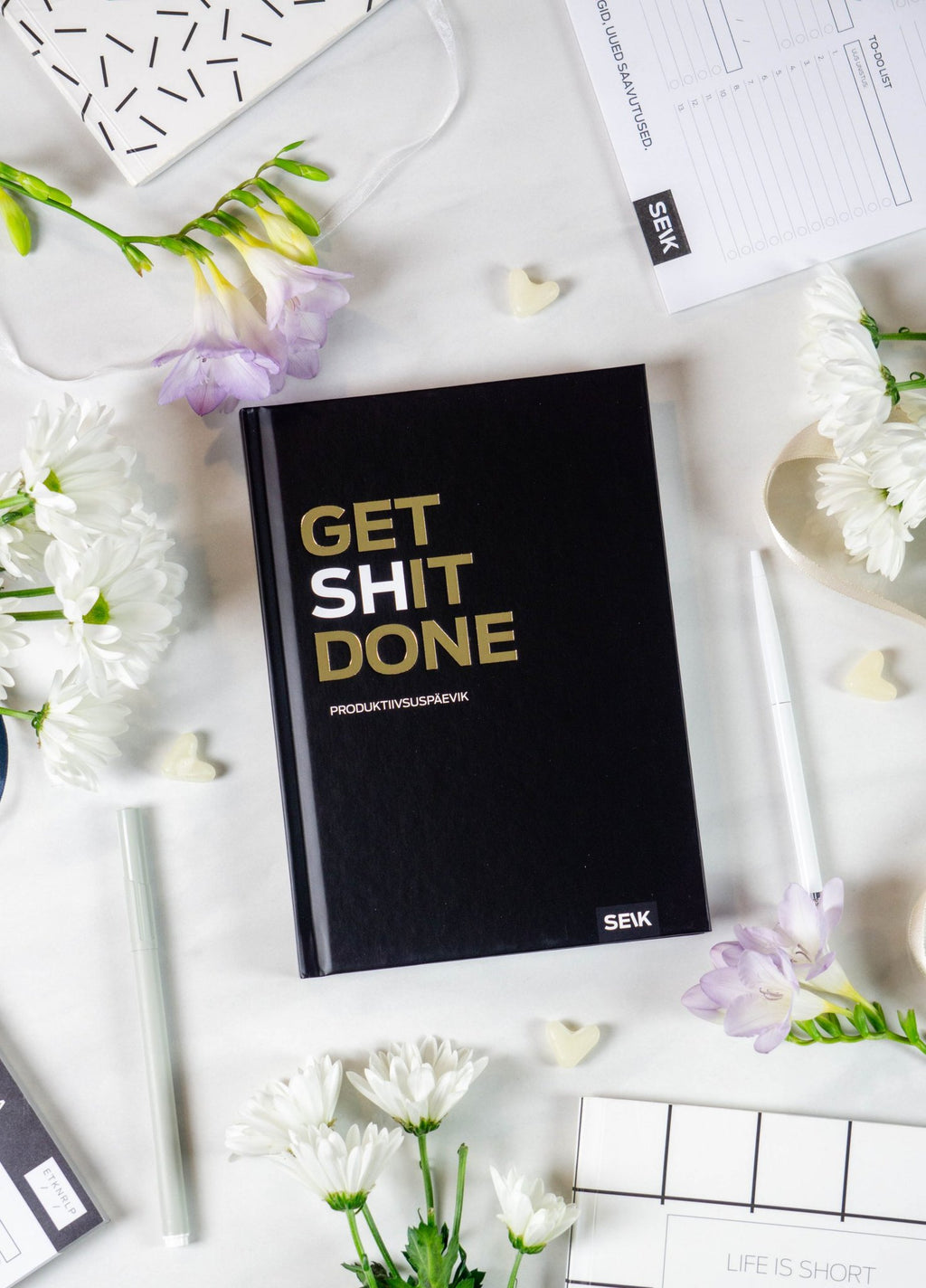 3. Cover of Get (sh)it done #Produktiivsuspäevik planner by SEIK, black with gold text, on a desk with flowers