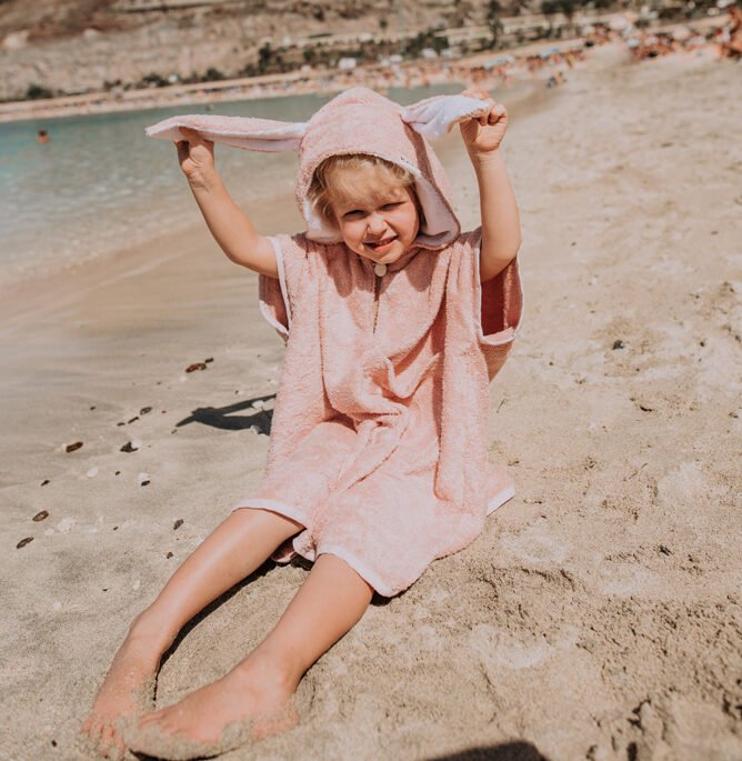 9. Close-up of toddler in pink RÄTT short-sleeve poncho sitting on the beach, holding hood ears
