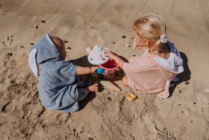 1. Kids wearing RÄTT short-sleeve ponchos in pink and blue, playing on the beach