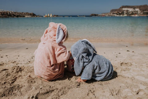 2. Back view of children in RÄTT short-sleeve ponchos, pink and blue, sitting on the beach