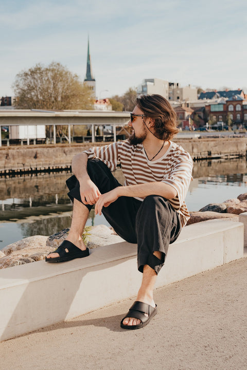 1. Men's black Miku sandals by Omaking worn by model sitting outdoors by the water