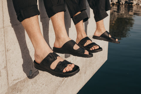 1. Men's black Märt Sandals by Omaking worn by two models sitting on a ledge, showcasing adjustable buckle straps